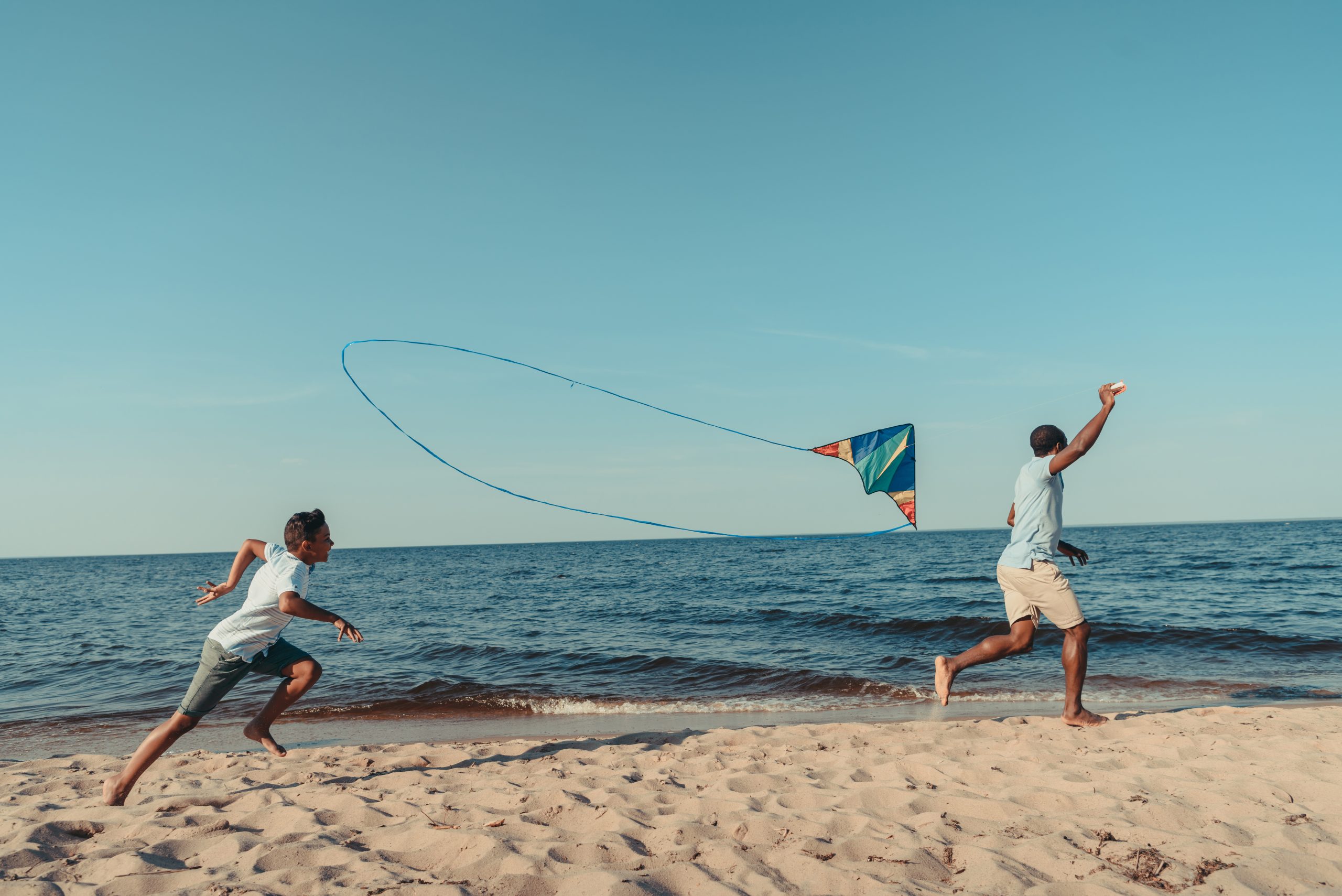 happy african american father and son playing with kite and running on sandy beach